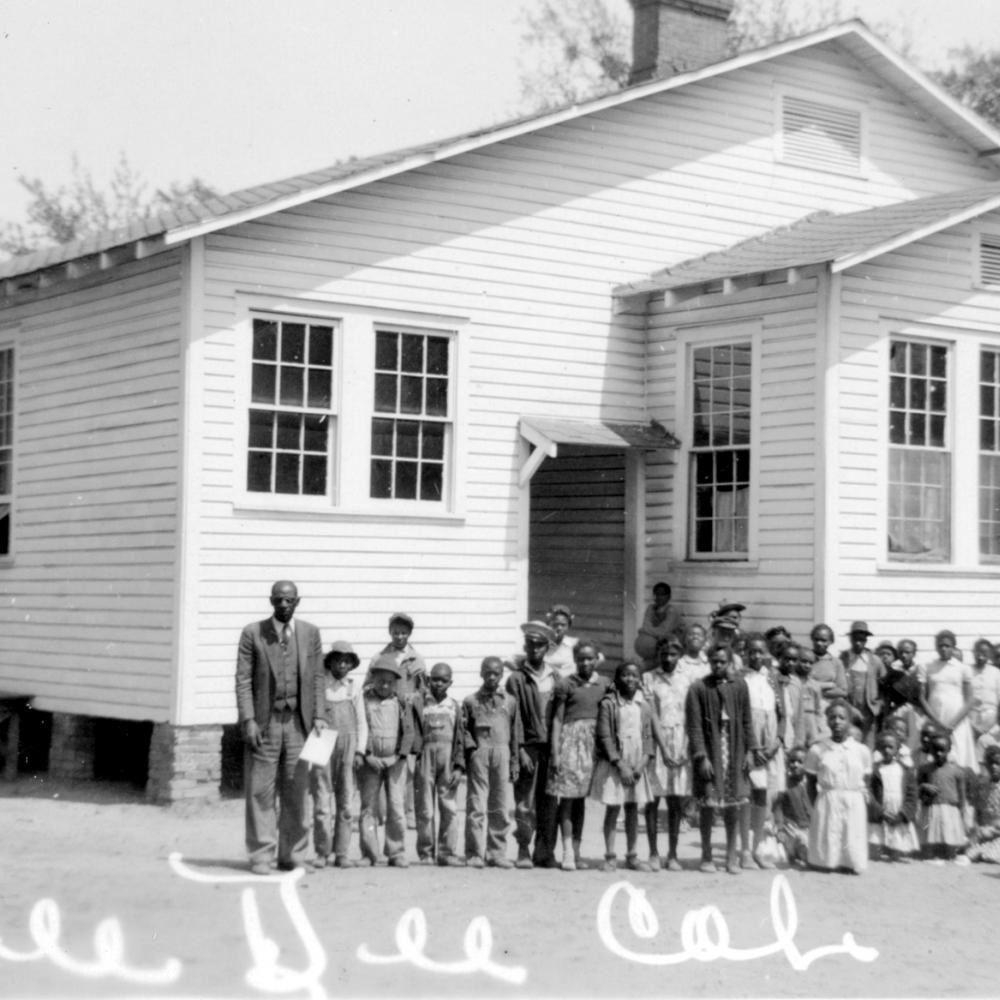Black and white photo of African American students standing in front of a small white schoolhouse.