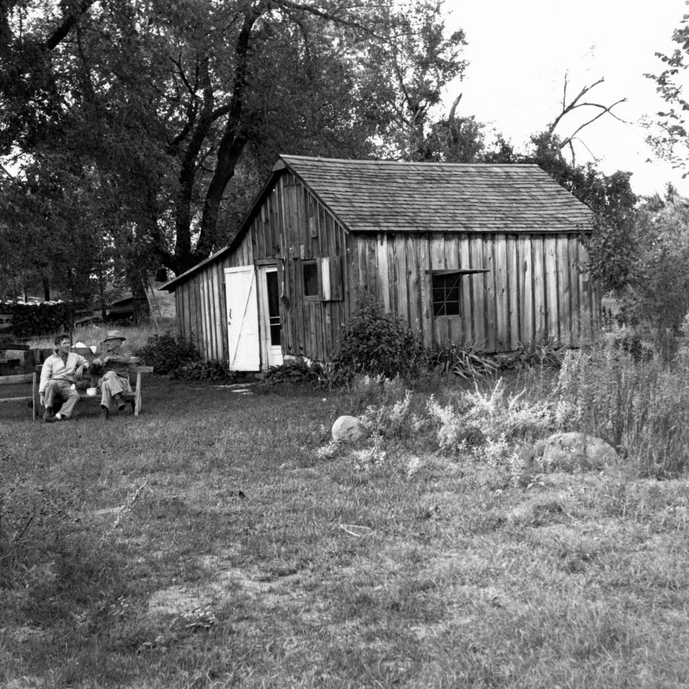 Black and white photo of two men on a bench outside a shack