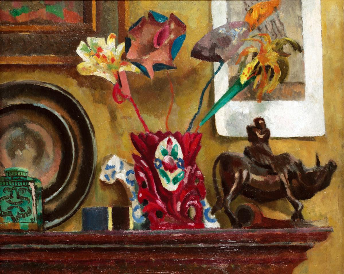 Brightly colored paper flowers in a red vase, set on a dark wood mantelpiece, surrounded by other small knickknacks and framed paintings