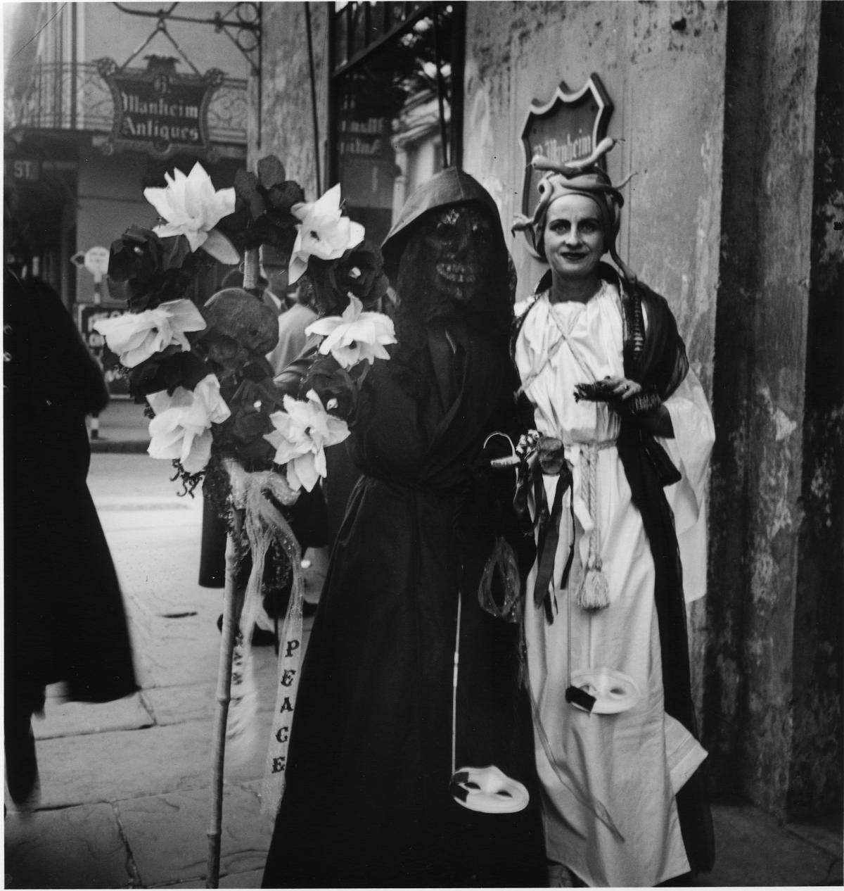 Two revelers stand on a New Orleans street; one wearing a long white robe and snake-topped cap, the other in a hooded black robe, black skull mask, and holding a wreath of black and white flowers