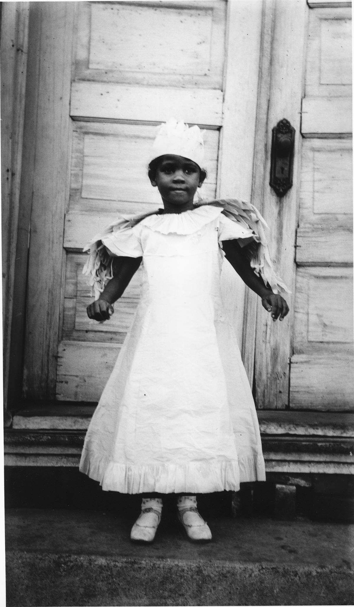 A young African American girl wears a white dress with wings sewn into the shoulders and a feathered flat cap