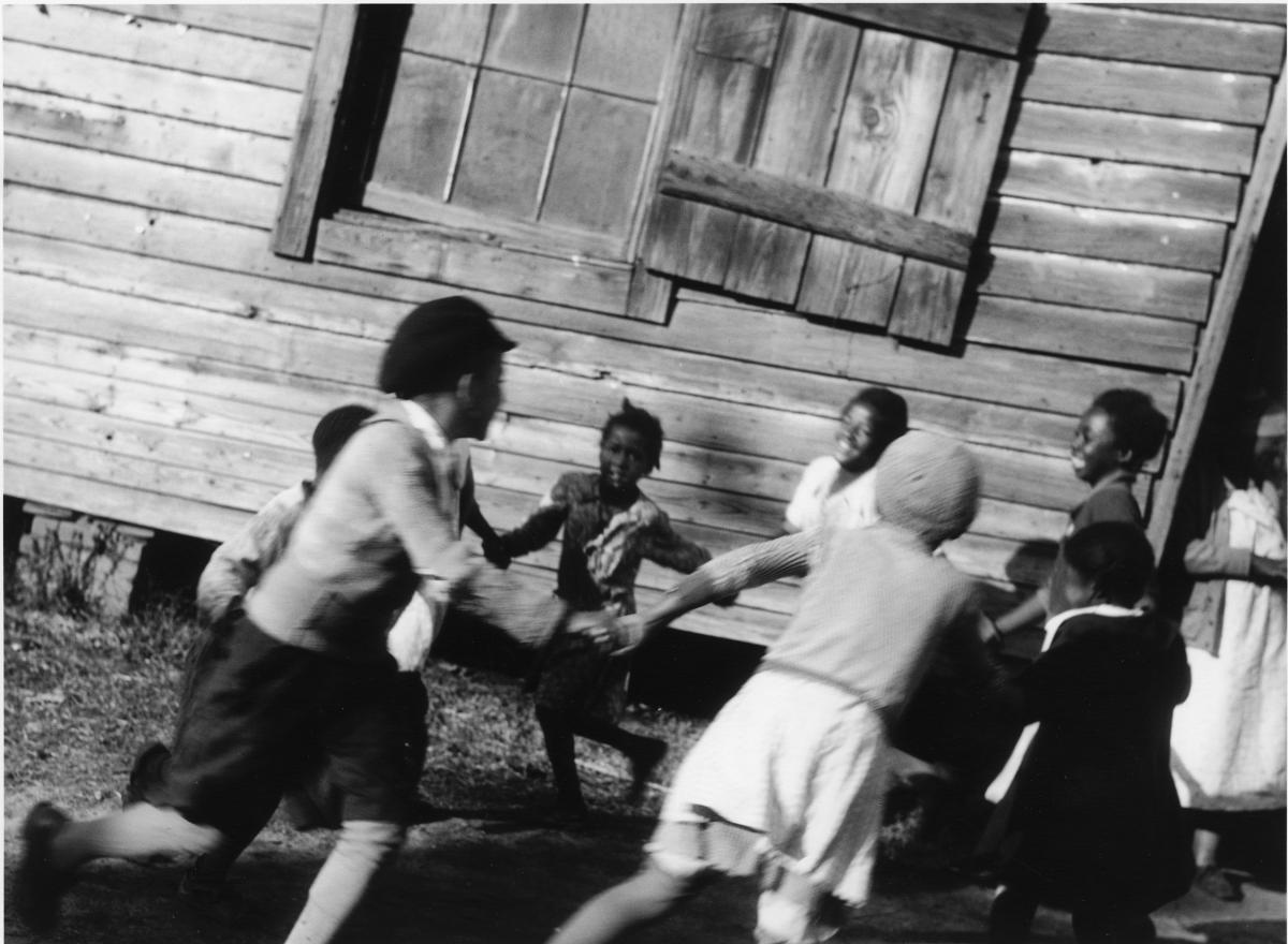 A group of schoolchildren link arms and run around in a circle in front of a wooden building