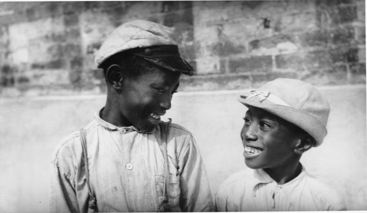 a pair of African American boys, both wearing caps, smile and look at each other
