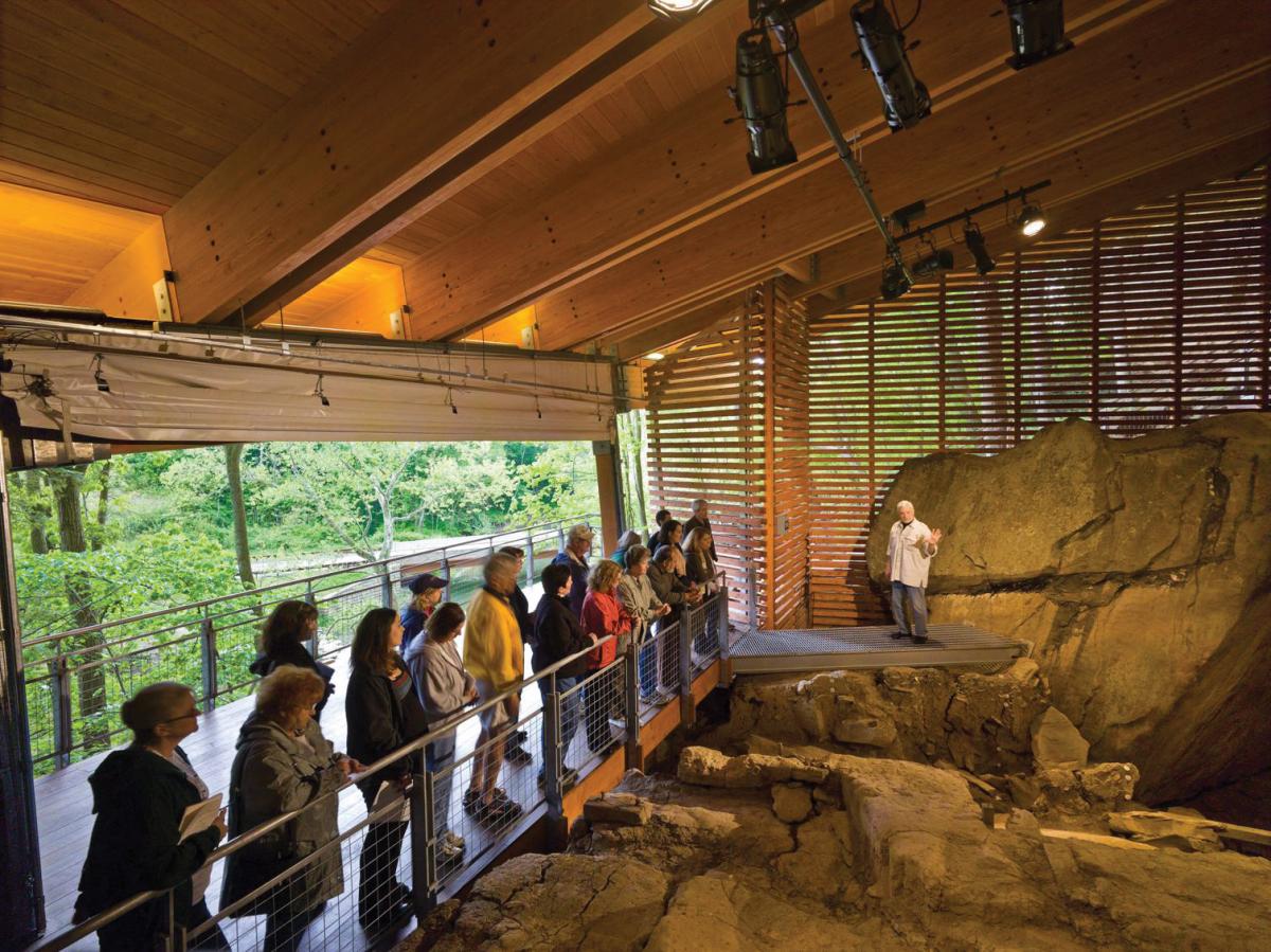 Visitors line up at a railing as Adovasio gestures to the rocky excavation site