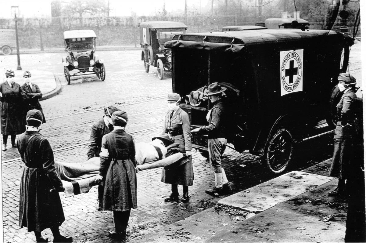Four masked volunteers carry a covered body on a gurney into a Red Cross truck, while masked officials look on from the sidewalk