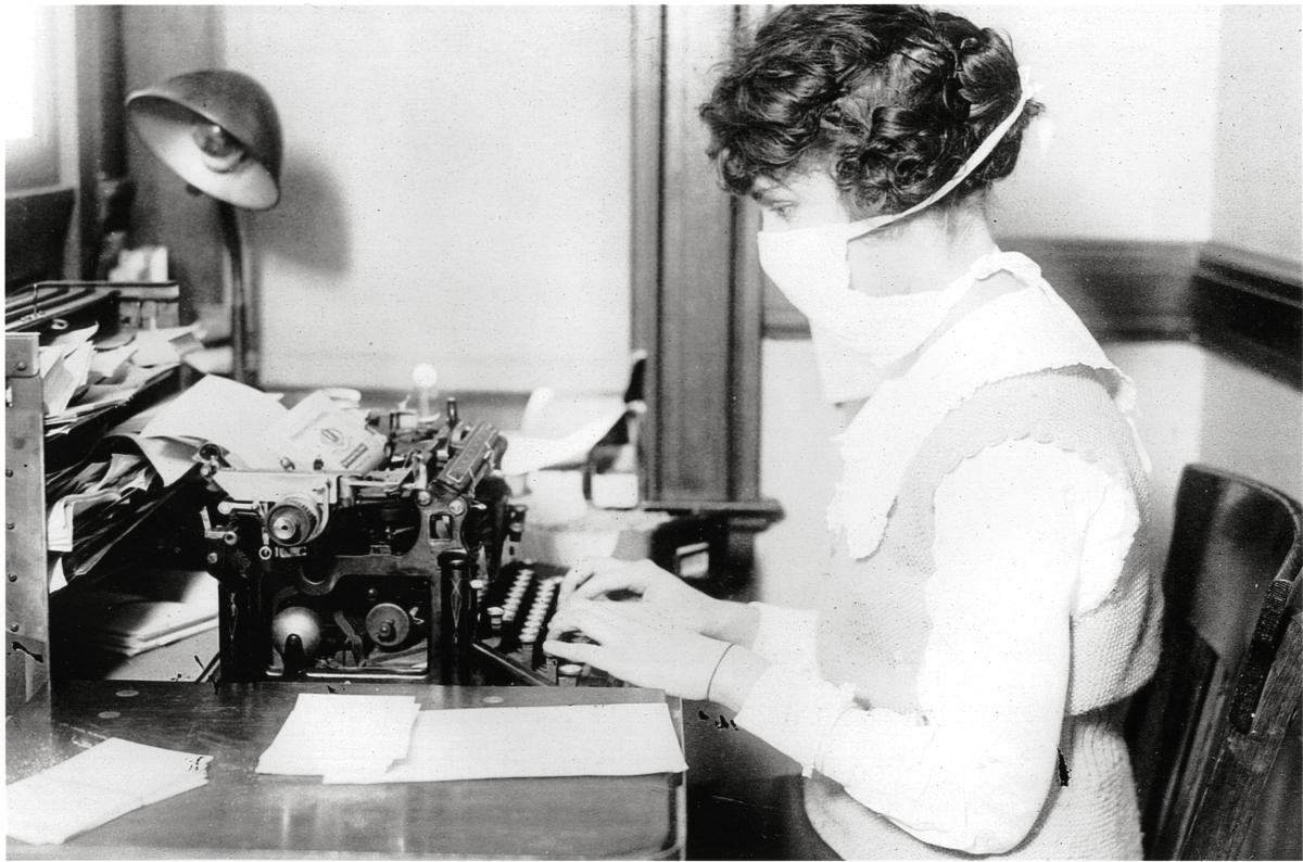 A woman types on a typewriter while wearing a thick white flu mask