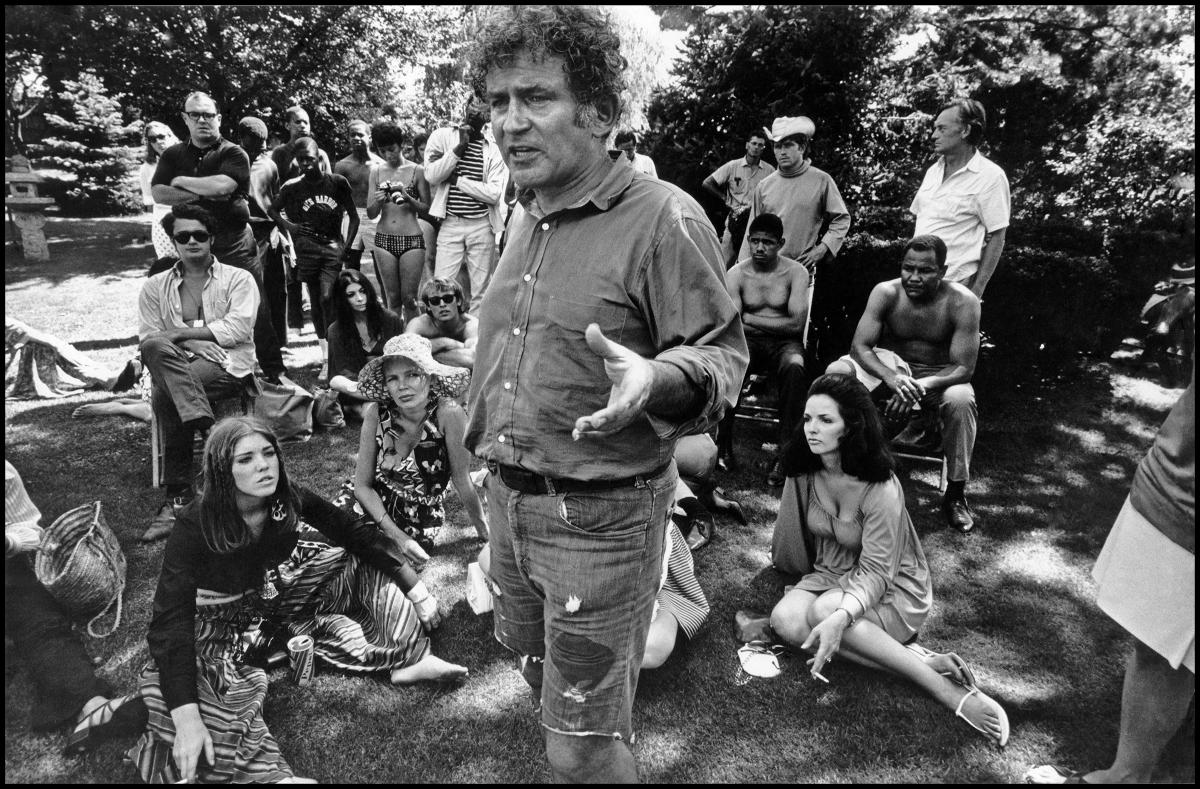 a white man stands and talks to a crowd of hippies