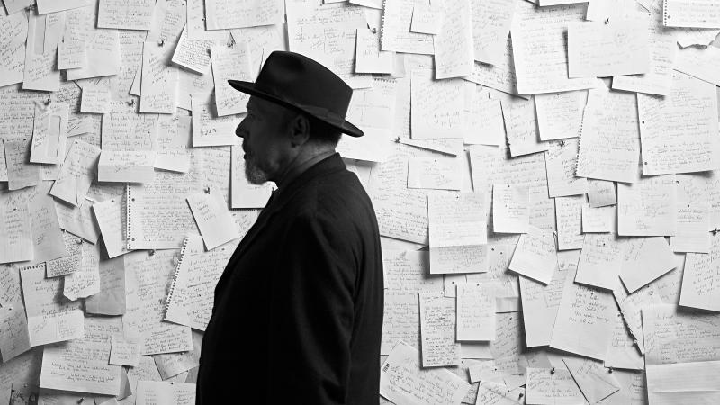 Black and white side portrait of August Wilson standing before a wall covered by letters that have been pinned onto it.