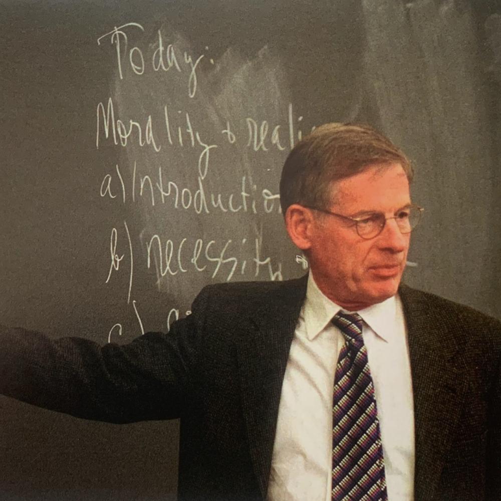 Mansfield lectures to a class at Harvard University. He is pointing at a green chalkboard in a black suit, patterned tie, white shirt, and glasses. He is facing the class.