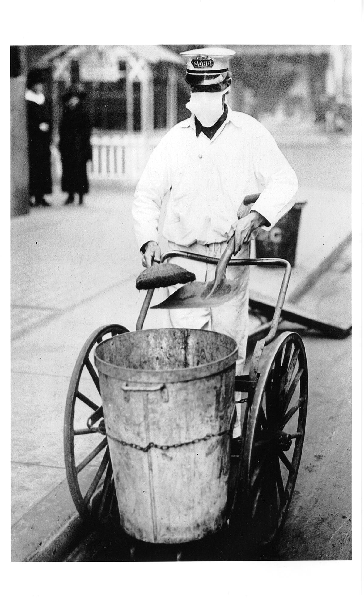 A white uniformed sweeper wearing a cloth mask that covers his entire face except the eyes, pushes his wheeled garbage can and sweeping supplies down the street