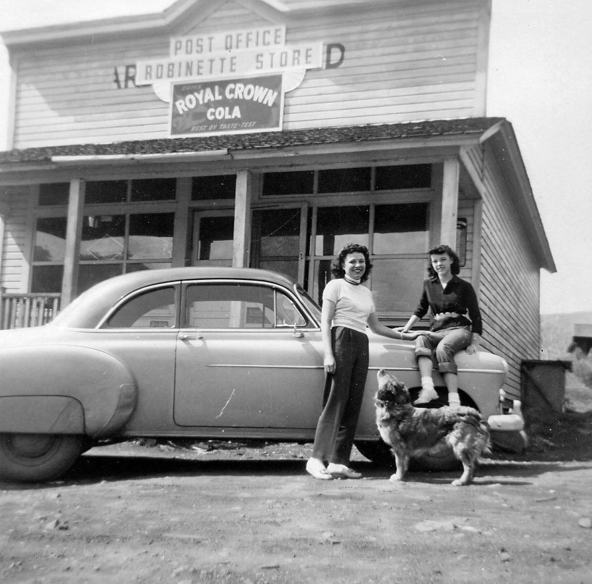 Black and white photo of a mother and her teenage daughter posing in front of the Robinette Post Office with their dog. 