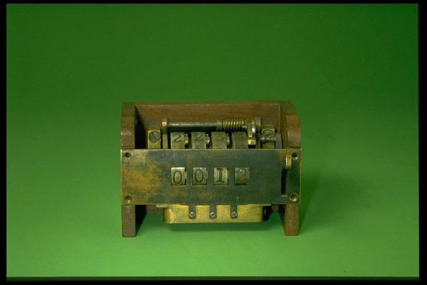 A small brass, steel, and wood counting machine with four digits on display sits on a green background.