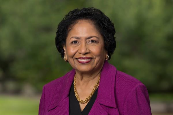 Dr. Ruth J. Simmons in a purple jacket and gold chain against a dark, leafy background