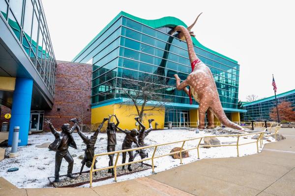 Image of the Indianapolis Children's Museum, featuring a colorful glass building, a statue of children playing, and statues of dinosaurs.