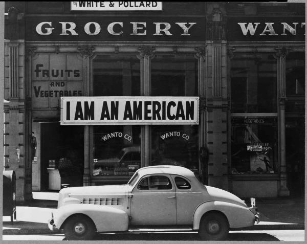 Black and white photo of a car in front of the storefront of a Japanese business that bears a sign reading "I am an American." 