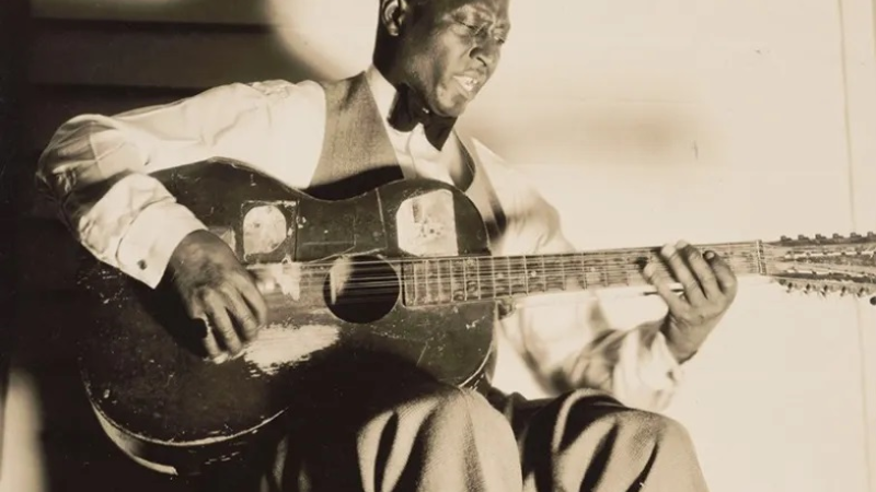 Black-and-white photo of the singer Lead Belly singing and playing the guitar. 