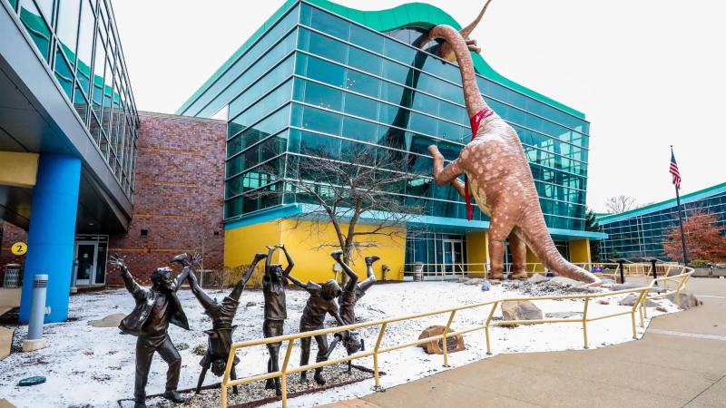 Image of the Indianapolis Children's Museum, featuring a colorful glass building, a statue of children playing, and statues of dinosaurs.